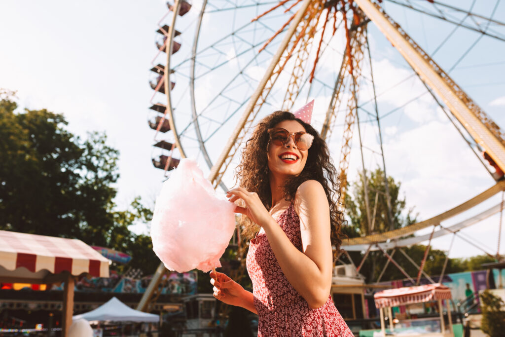 Smiling woman with dark curly hair in sunglasses and birthday cap standing with cotton candy in hand and happily looking aside while spending time in amusement park with ferris wheel on background